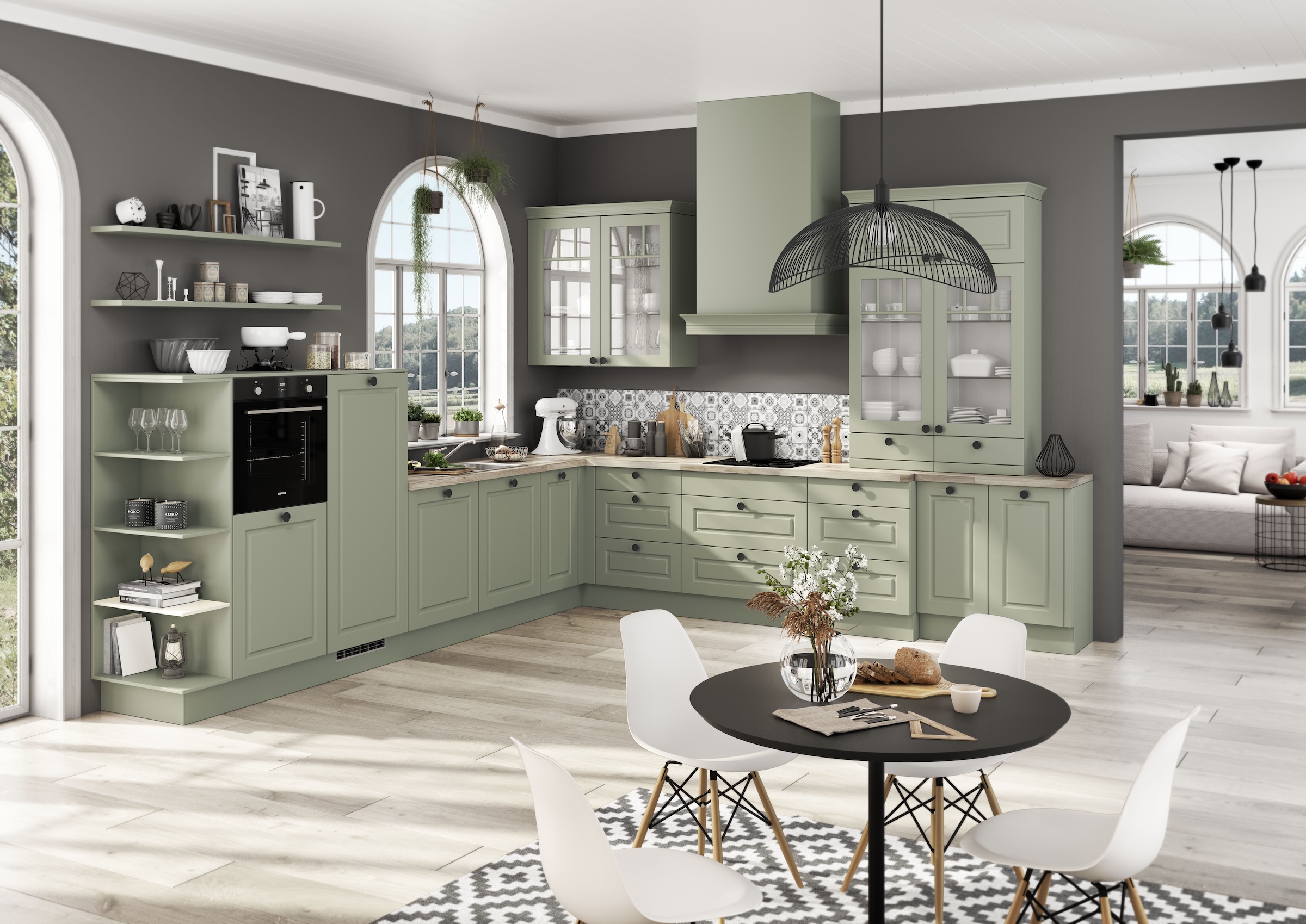 Sage-green kitchen with lower and wall cabinets, open shelving, and a round black dining table in a bright open-plan space with arched windows.