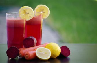 Two tall glasses of red juice with lemon slices, surrounded by lemons, carrots, and beets on a table nearby.