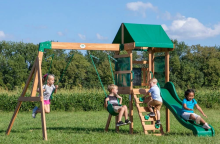 Four children play on a wooden playground with swings, a climbing wall and a green slide in a sunny field.