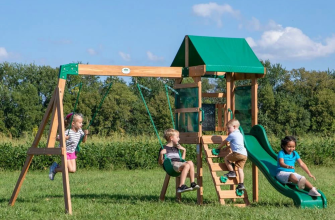 Four children play on a wooden playground with swings, a climbing wall and a green slide in a sunny field.