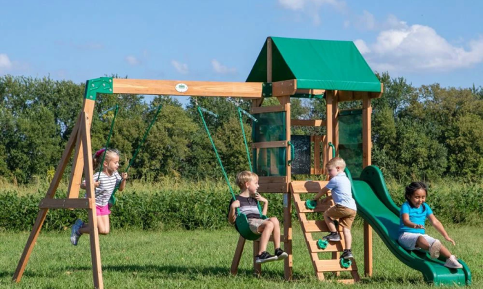 Four children play on a wooden playground with swings, a climbing wall and a green slide in a sunny field.