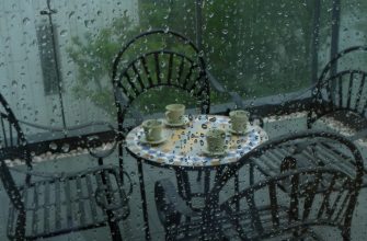Rainy patio with a round metal table and four chairs; several cups on the table, viewed through raindrop-covered glass.
