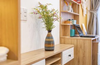 Wooden desk with a striped ceramic vase of pink and white flowers, beside a laptop on a shelf unit in a sunny room.