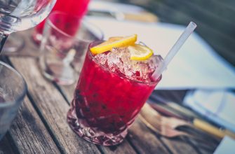 Red cocktail over ice with a lemon twisted garnish and straw on a rustic wooden table outdoors.