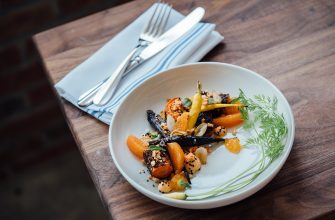White plate with a colorful roasted-vegetable dish: orange segments, onions, almonds, seeds, and fresh greens on a wooden table.