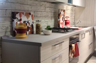 Modern kitchen counter with a yellow-red spice grinder, a jar of mixed toppings, a white bowl with a lime, and a gas cooktop against a white stone backsplash.