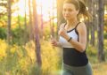 Woman jogging through a sunlit forest, wearing headphones and holding a smartphone.