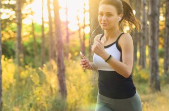 Woman jogging through a sunlit forest, wearing headphones and holding a smartphone.