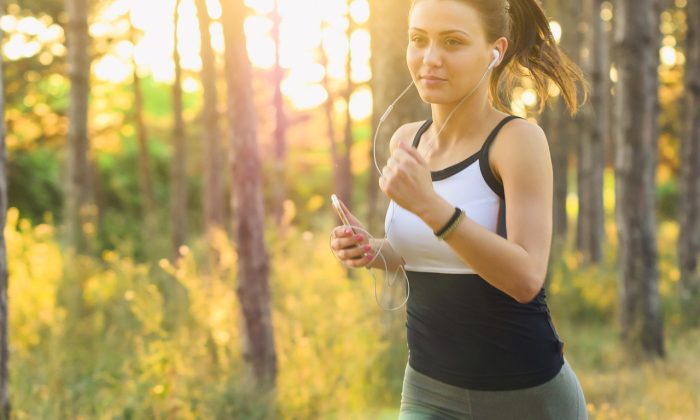 Woman jogging through a sunlit forest, wearing headphones and holding a smartphone.