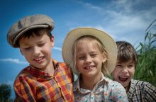 Three children outdoors on a sunny day wearing hats, smiling at the camera with blue sky behind them.