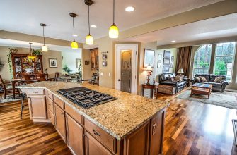 Open-concept kitchen and living room with a granite island, gas cooktop, pendant lights, and hardwood floors leading to a sofa area by a large arched window.