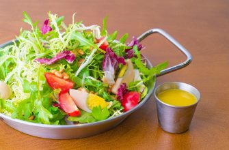 Colorful mixed salad in a metal pan with greens, tomato, egg, and nuts, plus a small cup of yellow dressing nearby.