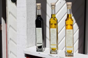 Three tall glass bottles of oil and vinegar lined up on a white ledge outside, two golden olive oils and a dark balsamic vinegar with labeled fronts visible