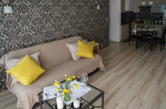 Beige sofa with yellow cushions in a living room with black damask wallpaper and a glass coffee table with daffodils and candles in the foreground