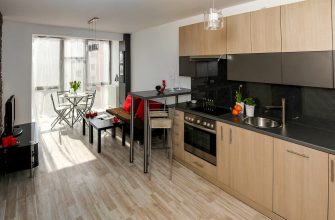 Open-plan kitchen with light-wood cabinets, black countertop, stainless oven, and a small island/bar beside a red-cushioned sofa area.