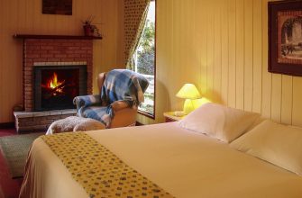 Cozy bedroom with a lit fireplace, king-size bed, and a chair by a window covered with a checkered blanket.