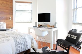 Bedroom with a white desk and iMac, a modern chair, and a bed with graphic bedding; large windows and wood accent wall nearby.