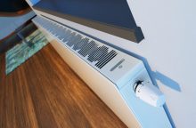Baseboard heater with vent grille and a round thermostat knob mounted along a wall above a wooden floor; green indicator lights are on.