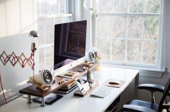 Home office desk with an iMac on a wooden stand, speakers, keyboard, and organized supplies by a window.