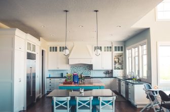 Bright white kitchen with a blue island, pendant lights, and large windows on the right, featuring stainless appliances.